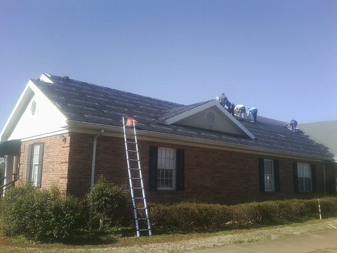 Crew tearing shingles off a brick ranch roof with a ladder set — Metal Roof Repair work by our crew in Dannemora