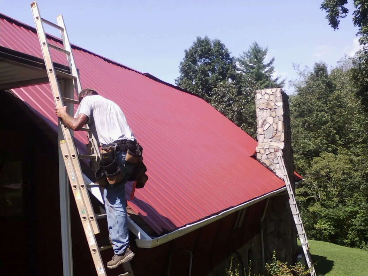 Roofer on a ladder beside a red metal roof and stone chimney — Soffit & Fascia Repair by Roofing Brothers in Dannemora